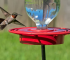 Hummingbird approaches a vibrant red hummingbird feeder against green grass.